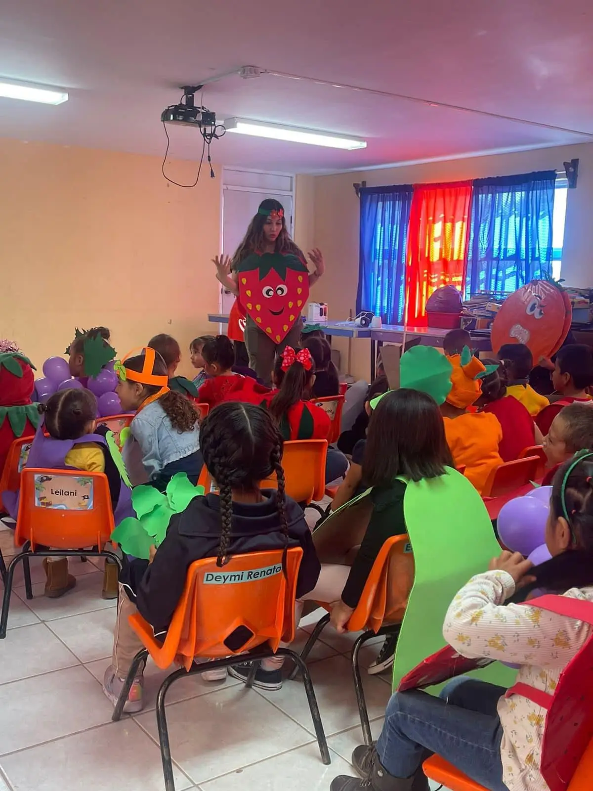 Reyna Isabel Gutierrez Pierce disfrazada de una fruta dando una clase para niños en una primaria. La clase esta llena y todos los niños estan disfrazados de frutas y verduras.